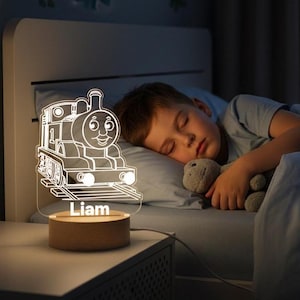Könnte beinhalten: Ein Nachtlicht mit einem Cartoon-Zug-Design, das den Namen "Liam" auf seinem Holzsockel anzeigt. Das klare, geätzte Zugbild ist beleuchtet. Die Lampe steht auf einem Nachttisch neben einem schlafenden Kind.