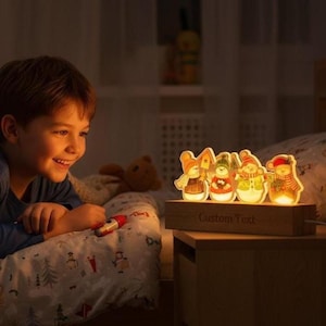 May include: A wooden night light with four illuminated snowman figures. The base of the light has the text "Custom Text". The snowmen are wearing hats and scarves. The light is on a wooden nightstand.