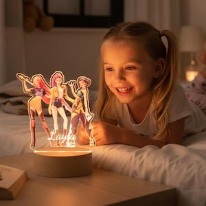 Peut inclure: Une veilleuse LED décorative avec trois personnages stylisés aux tenues colorées. La lumière est montée sur une base en bois ronde et porte le nom "Layla" illuminé à l'avant. Une jeune fille sourit à la lumière.