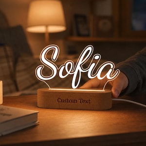 Pode incluir: Uma luz noturna LED personalizada com o nome Sofia em letras cursivas brancas num painel de acrílico transparente. O painel está montado numa base de madeira lisa e oval com as palavras Custom Text gravadas na parte frontal da madeira.