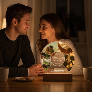 May include: A personalized photo lamp with a wooden base and a circular acrylic panel. The panel displays multiple photos, a date, song information, and a music waveform. The lamp is illuminated, creating a warm glow.
