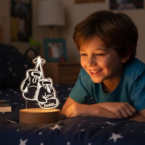 May include: A decorative night light featuring a pair of boxing gloves. The gloves are white with black outlines and the name "DANIEL" is printed on one of the gloves. The light sits on a wooden base and is illuminated.