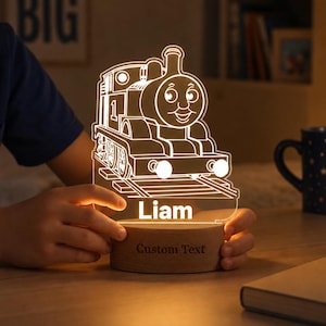 May include: A custom LED night light featuring an acrylic cutout of a steam locomotive train with the number one on its side. The name Liam is etched below the train. The light sits on a round wooden base with the text Custom Text engraved on it.