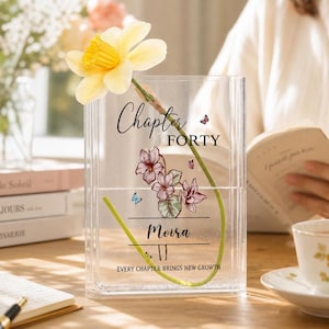 Peut inclure: Un vase rectangulaire transparent en acrylique contenant une tige de jonquille jaune. La face avant porte l'inscription "Chapter FORTY", le prénom "Moira" et "EVERY CHAPTER BRINGS NEW GROWTH", avec des illustrations de fleurs violettes et de papillons.