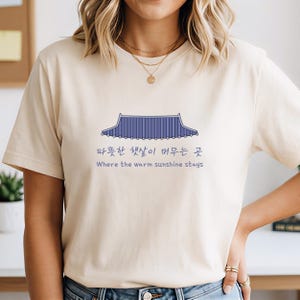May include: A cream-coloured t-shirt with a blue illustration of a Korean roof design. The text below the graphic, in Korean and English, reads "Where the warm sunshine stays."