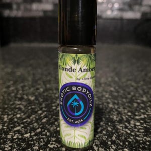 May include: A small glass bottle with a black cap and a green, white, and purple label. The label says "Blonde Amber by: Clive Christian Exotic Body Oilz Est. 2014".