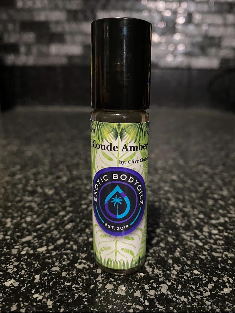 May include: A small glass bottle with a black cap and a green, white, and purple label. The label says "Blonde Amber by: Clive Christian Exotic Body Oilz Est. 2014".