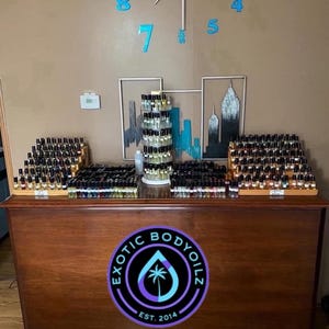 May include: A wooden counter with a variety of small glass bottles filled with liquid. The counter has a sign that says "Exotic Body Oils Est. 2014".