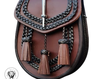 Puede incluir: Un sporran de cuero marrón con correas trenzadas negras y borlas. El sporran tiene un broche de kilt plateado con un diseño decorativo.