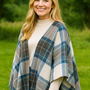 May include: A woman wearing a blue, brown, and white plaid cape with a cream-colored turtleneck. The cape has a fringed edge. The background is a blurred green outdoor scene.