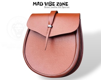 Può includere: Un sporran in pelle marrone con chiusura a patta e cinturino. Lo sporran ha un fondo arrotondato e una superficie strutturata. Il testo "MAD VIBE ZONE Brown Leather Kilt Sporran" è in alto.