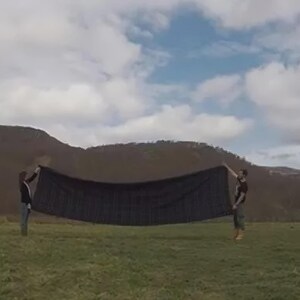 Pu&ograve; includere: Due persone tengono in mano un grande oggetto rettangolare nero all'aperto. L'oggetto sembra essere un tessuto o un telone, teso contro uno sfondo di erba verde, dolci colline e un cielo nuvoloso.