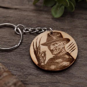 Op de afbeelding: Een ronde houten sleutelhanger met een donker, lasergegraveerd portret van een personage met een fedora-hoed en een handschoen met lange, scherpe messen aan de vingers. De sleutelhanger is bevestigd aan een standaard zilverkleurige metalen ring.