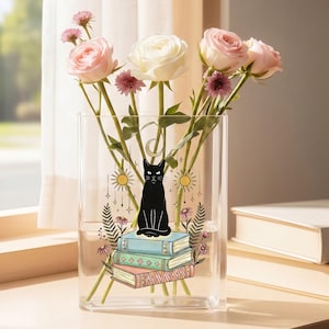 Pode incluir: Um vaso retangular de vidro transparente com uma ilustração de um gato preto sentado sobre uma pilha de três livros, rodeado por símbolos de sol e lua. O vaso contém várias rosas cor-de-rosa claras e brancas com caules verdes e pequenas flores roxas.