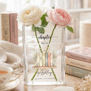 May include: A clear rectangular glass vase featuring a printed graphic of a bookshelf with books, flowers, and butterflies. The text reads A NEW CHAPTER BEGINS, Ashley, and YOUR STORY IS JUST BEGINNING. Two large roses, one white and one pink, are inside.