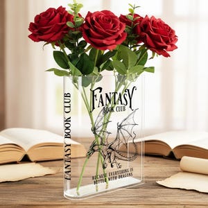Peut inclure: Un vase transparent en acrylique en forme de livre, avec le texte "FANTASY BOOK CLUB" et une illustration de dragon. Il contient des roses rouges. Le texte "BECAUSE EVERYTHING IS BETTER WITH DRAGONS" est également visible.