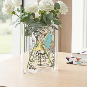 May include: A clear glass vase with a Beauty and the Beast graphic, holding white roses and greenery. The graphic features Belle and the Beast, with text that reads "Beauty and the Beast" and "Bold as a Rose". The vase sits on a light-colored wooden surface.