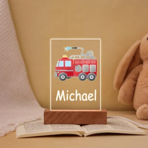 Pode incluir: Luz noturna personalizada com um design de caminhão de bombeiros vermelho e o nome "Michael" em branco. A luz está em uma base de madeira. O fundo é bege claro.