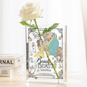 Può includere: Un vaso trasparente in acrilico con un design di "La Bella e la Bestia", con Belle e la Bestia. Il vaso contiene una rosa bianca. Il testo sul vaso recita "Beauty and the Beast" e "Enchanted Beauty".