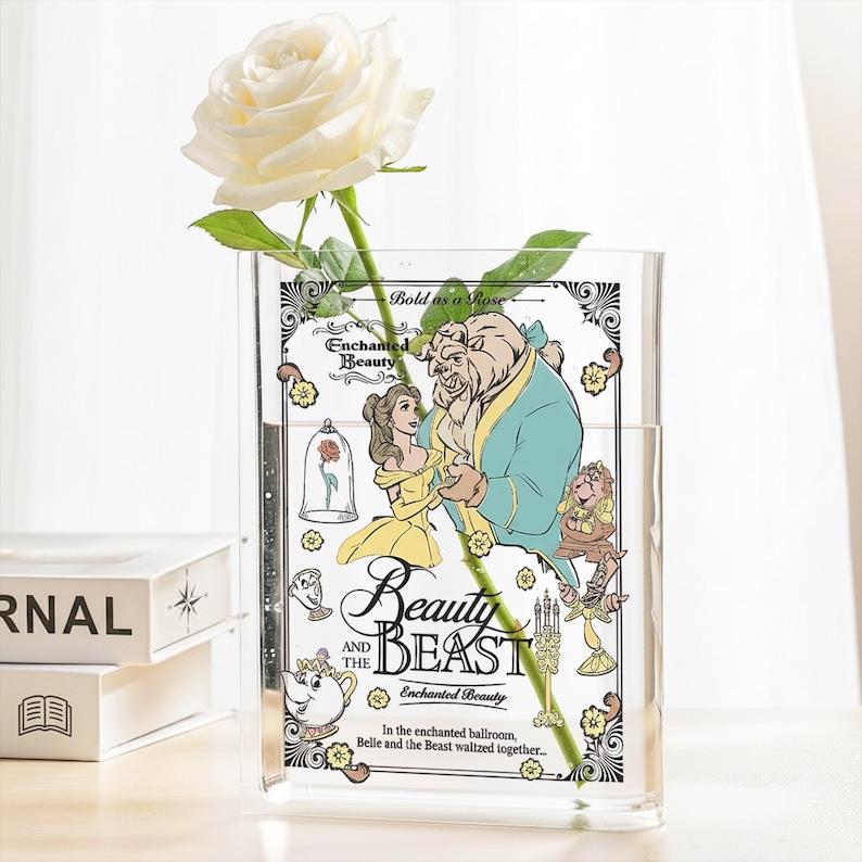 May include: A clear acrylic vase featuring a "Beauty and the Beast" design with Belle and the Beast. The vase holds a white rose. Text on the vase reads "Beauty and the Beast" and "Enchanted Beauty".