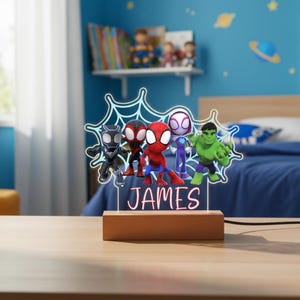 May include: A wooden base night light featuring superhero figures, including Spider-Man and Hulk, with the name "JAMES" illuminated in pink. The figures are set against a blue spiderweb design.