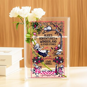Può includere: Vaso in acrilico trasparente con la copertina del libro "Alice's Adventures in Wonderland & Other Stories". Il vaso contiene rose bianche. La copertina presenta illustrazioni di Alice, un coniglio bianco e un gatto.