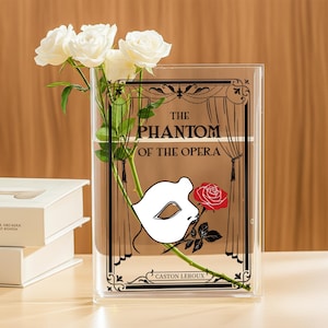 May include: Clear rectangular vase with a graphic from "The Phantom of the Opera" featuring a white mask and red rose. White roses are arranged inside the vase. The vase is on a light-colored surface.