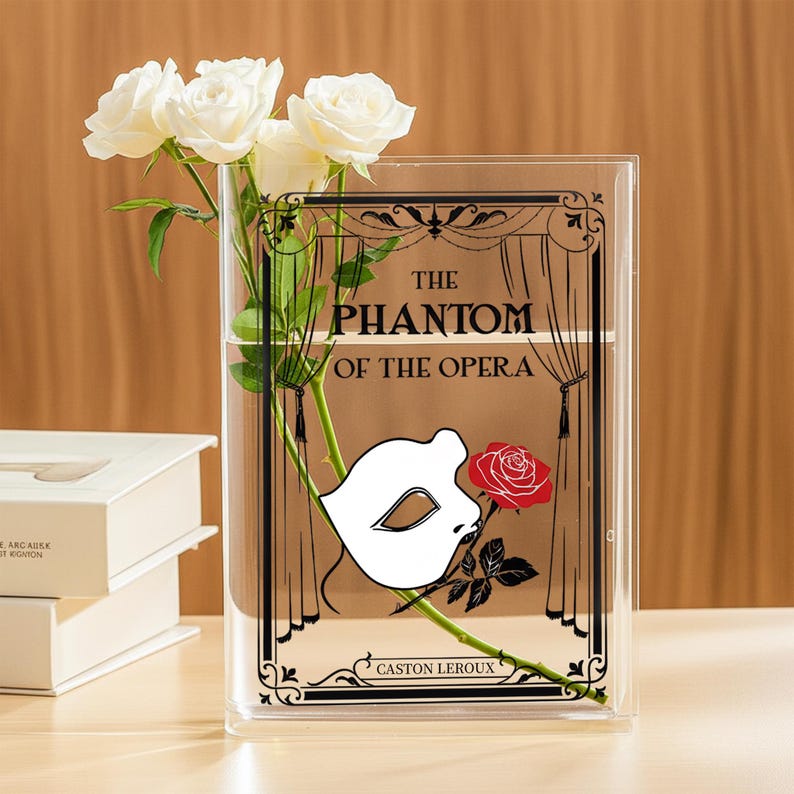 May include: Clear rectangular vase with a graphic from "The Phantom of the Opera" featuring a white mask and red rose. White roses are arranged inside the vase. The vase is on a light-colored surface.