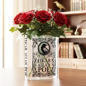 May include: A clear glass vase shaped like a book, featuring the text "EDGAR ALLAN POE" and a raven illustration. The vase holds a bouquet of vibrant red roses, green ivy, and white filler flowers. The vase is on a wooden table.