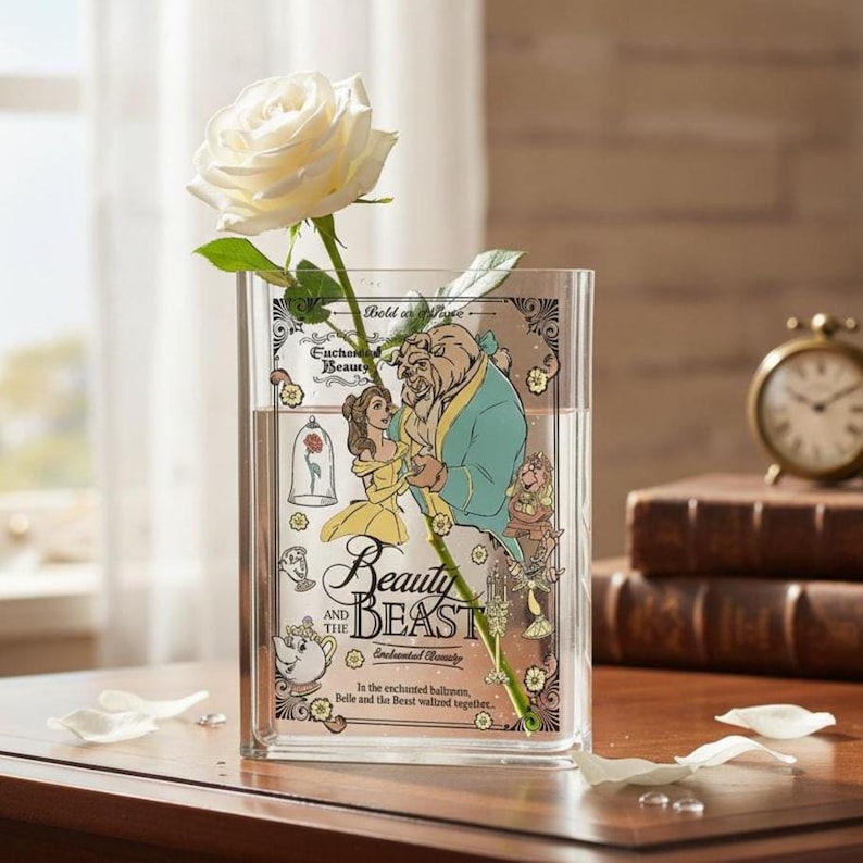 May include: A clear, rectangular vase featuring a "Beauty and the Beast" design, holding a white rose. The vase has a decorative illustration of Belle and the Beast. A small, gold-colored clock and books are in the background.