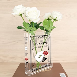 May include: Clear acrylic vase in the shape of a book, with the title "The Phantom of the Opera" and a rose design. The vase contains white roses and green foliage, placed on a brown surface.