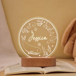 May include: A personalized night light with the name "Jessica" in elegant script. The circular design features sea creatures like a whale, turtle, and crab, illuminated by a warm glow from the wooden base. A charming decorative piece.