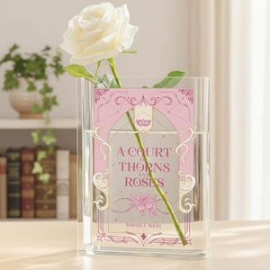 Puede incluir: Un jarrón de vidrio transparente con el texto "A Court of Thorns and Roses" sobre un fondo rosa. Una rosa blanca está colocada dentro del jarrón. El jarrón está sobre una superficie de madera.