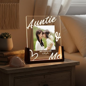 May include: Illuminated photo frame with the words "Auntie & Me" in a handwritten style. The frame displays a photograph of two people. The frame is lit with a warm glow and rests on a wooden bedside table.