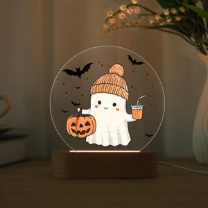 May include: A Halloween-themed night light featuring a cartoon ghost wearing an orange knit cap. The ghost holds a carved pumpkin and a drink with a straw. The design is on a clear, circular acrylic panel, set on a wooden base.