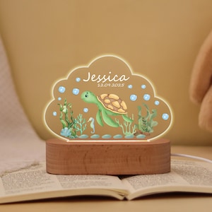Op de afbeelding: Decoratief nachtlampje met een zeeschildpadontwerp, de naam "Jessica" en de datum "12.09.2025". Het licht heeft een houten voet en een wolkvormige acryl bovenkant met een illustratie met zeethema.