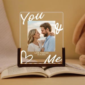 Op de afbeelding: Een verlichte acryl fotolijst met de woorden "You & Me" en een hartvormgeving. De lijst bevat een foto van een stel en de tekst "LOVE FOREVER 02.14.2026". De lijst staat op een houten voet.