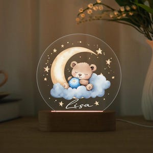 Peut inclure: Veilleuse avec un ours en peluche endormi sur un croissant de lune et un nuage. L'ours tient une balle bleue. Le nom "Lisa" est écrit en dessous. La lumière repose sur une base en bois.