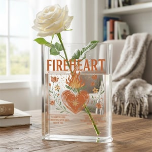 Könnte beinhalten: Transparente rechteckige Glasvase mit dem Wort "FIREHEART" in orangefarbenen Buchstaben. Die Vase hat ein orangefarbenes Herzdesign mit Flammen und floralen Akzenten. Eine weiße Rose befindet sich in der Vase.
