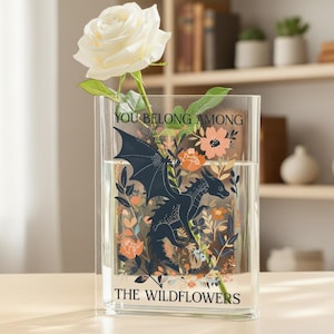 May include: A clear glass vase featuring a dragon and floral design, with the text "YOU BELONG AMONG THE WILDFLOWERS". A white rose is placed inside the vase.