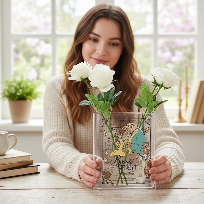 May include: Clear acrylic vase featuring a Beauty and the Beast design, holding white roses. The vase has a rectangular shape with a decorative illustration of Belle and the Beast. The roses have green stems and leaves.