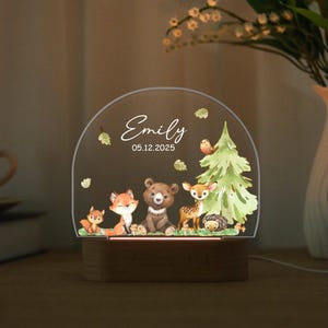 May include: A decorative night light featuring a woodland scene with a fox, bear, deer, and hedgehog. The light has a wooden base and a clear, arched top with the name "Emily" and the date 05.12.2025.