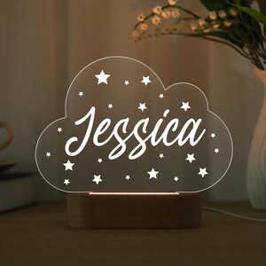 May include: A cloud-shaped night light with the name "Jessica" in white script. The clear acrylic light has star cutouts and sits on a wooden base. A warm glow emanates from the base, illuminating the name and stars.