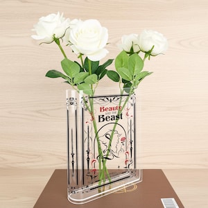 Può includere: Vaso trasparente in acrilico a forma di libro, con il titolo "Beauty and the Beast" e un'illustrazione dei personaggi. Il vaso contiene rose bianche e foglie verdi. Il vaso è su una superficie marrone.