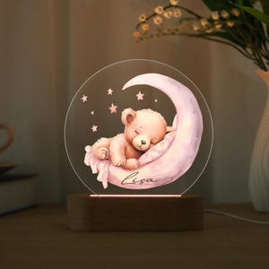 May include: A night light featuring a sleeping teddy bear on a pink crescent moon, surrounded by stars. The bear rests on a pillow, with the name "Lisa" written below. The light sits on a wooden base.