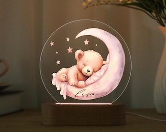 May include: A night light featuring a sleeping teddy bear on a pink crescent moon, surrounded by stars. The bear rests on a pillow, with the name "Lisa" written below. The light sits on a wooden base.