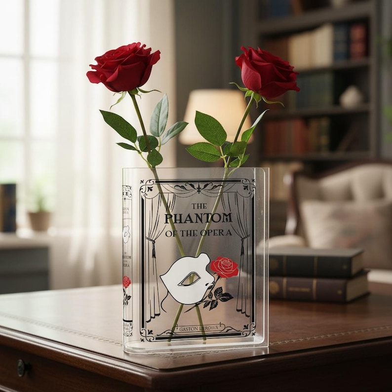 May include: Clear acrylic vase shaped like a book, featuring the text "The Phantom of the Opera" and a mask design. Two red roses with green leaves are placed inside the vase, creating a decorative floral arrangement.