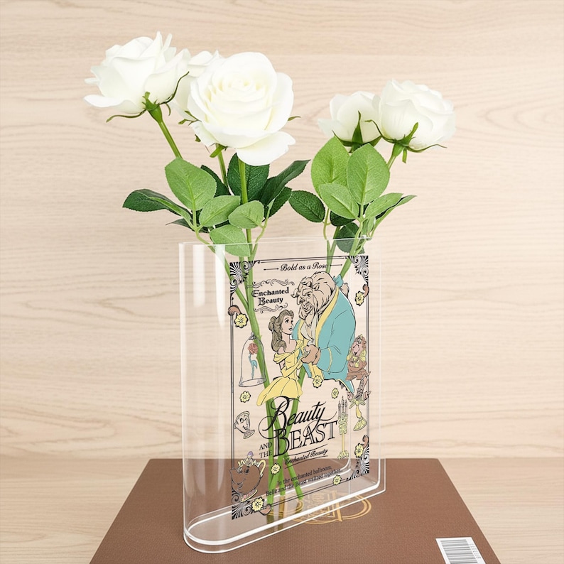 May include: A clear acrylic vase featuring a Beauty and the Beast design, holding white roses with green leaves. The vase has a curved shape and is set on a brown surface. The design includes the words "Beauty and the Beast".