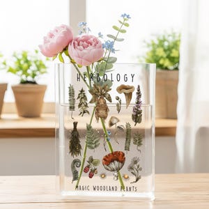 May include: A clear rectangular vase with the word "HERBOLOGY" and "MAGIC WOODLAND PLANTS" printed on it. The vase contains pink peonies, blue flowers, and green stems. The vase is decorated with illustrations of various plants and mushrooms.