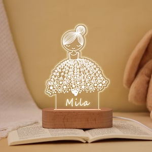 May include: A decorative night light featuring a ballerina design with the name "Mila" illuminated in white. The ballerina is wearing a floral dress, and the light sits on a wooden base. The light is connected to a white cord.